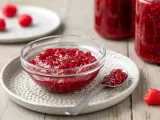 Una ciotola di vetro piena di marmellata di lamponi fatta in casa, con un cucchiaino e lamponi freschi sullo sfondo.