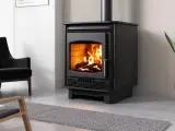 Poêle à bois moderne, flammes vives réchauffant la pièce. Les granulés ne tombent plus, juste du bois qui crépite. Un fauteuil noir et une plante verte complètent la scène.