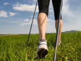 Gambe che praticano nordic walking tecnica su un prato verde sotto un cielo azzurro con nuvole bianche.