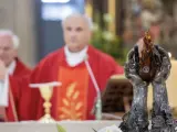 Dwie dłonie trzymają płomień. W tle ksiądz w czerwonej szacie, jak siostra zakonna, błogosławi wiernych.