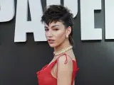 Mujer con un llamativo mullet largo, maquillaje rojo y vestido escarlata, posando ante un fondo con letras blancas.