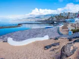 Słoneczna plaża w Playa de las Américas z basenem, łódką i kolorowymi budynkami w tle.