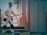 Man i arbetskläder använder en lång roller för att måla huset. En hink med färg står på gräset.