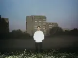 Bartosz Kruczyński stoi na tle bloków mieszkalnych, otoczony mgłą i kwitnącymi kwiatami.