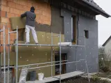 Pracownik na rusztowaniu montuje wełnę mineralną na ścianie domu. Brak cokołu na elewacji, trwa ocieplanie.