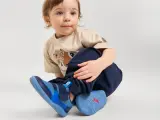 Małe dziecko w granatowych spodniach i beżowej koszulce z jelonkiem, na nogach niebieskie buty. Jaki to rozmiar buta dziecka? 26.