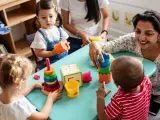 Zwei Frauen und drei Kleinkinder spielen mit bunten Spielsachen. Eine Frau hilft einem Kind, Türme zu stapeln, was eine tolle Methode ist, um Kinder zweisprachig zu erziehen.