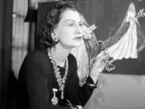 Coco Chanel, icona di stile, con occhiali in mano, sullo sfondo schizzi di abiti da sposa. Un'immagine che racconta la sua storia.