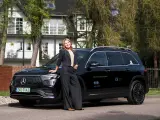 Kobieta w czarnym garniturze opiera się o czarnego Mercedesa EQB na tle białego domu.