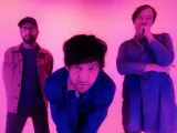 Trois hommes posent devant un fond rose. L'homme du milieu, penché en avant, a des yeux brillants et un regard intense.