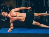 Un homme musclé réalise un gainage planche sur un tapis bleu dans une salle de sport.