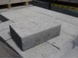 Stos szarych bloczków betonowych, każdy z widocznymi nierównościami i kruszywem. Wymiary bloczków betonowych są standardowe dla budowy.