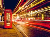 Klasyczna czerwona budka telefoniczna na tle nocnej ulicy, gdzie długi czas naswietlania zdjecia uchwycił smugi świateł samochodowych.