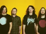 Quatre hommes aux cheveux longs posent devant un fond jaune vif. Leurs t-shirts portent des graphismes distinctifs, évoquant l'énergie brute du magma et la puissance de Gojira.