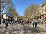 Tłum ludzi spaceruje po słonecznej La Rambla w Barcelonie, otoczonej drzewami i zabytkowymi budynkami.
