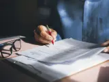 Persona revisa y firma una nota de encargo inmobiliaria, con gafas y bolígrafo rojo sobre documentos.