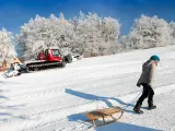 Dziecko ciągnie sanki po ośnieżonym stoku, szukając idealnego miejsca gdzie na sanki w góry. W tle czerwony ratrak przygotowuje trasę.
