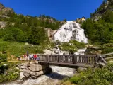 Cascata imponente con escursionisti su un sentiero, un ponte di legno attraversa un torrente. Un esempio di cosa vedere in Piemonte per gli amanti della natura.
