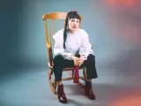 Scout Niblett, assise sur un fauteuil à bascule, porte une blouse blanche à volants et un pantalon noir. Elle tient un pompon rouge.