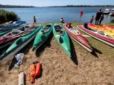 Kajaki czekają nad jeziorem. Kilka osób przygotowuje się do wodnych przygód, a w tle widać łodzie i żaglówki.
