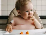 Małe dziecko w różowym śliniaku próbuje kawałek marchewki. Na tacce leżą kolejne pomarańczowe kawałki.