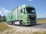 Zielona ciężarówka MAN TGX z naczepą TIP jedzie drogą. Na naczepie widnieje napis "Ucz się z najlepszymi!".