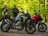 Dwóch motocyklistów z kaskiem na siedzeniu obok dwóch motocykli Ducati Multistrada, szary i czerwony, na tle leśnej drogi.