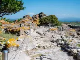 Ancient Italian ruins with weathered stones covered in orange lichen, a pathway winding through the site, and a distant blue sea under a clear sky.