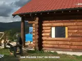 Une maison en rondins de bois en construction, avec un homme travaillant à proximité. Le toit est rouge et le paysage est montagneux.