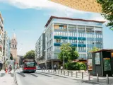 Un bus rouge circule dans une rue bordée d'immeubles et d'un clocher, sous un ciel bleu.