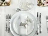 Ett elegant dukat bord med vita tallrikar, servetter dekorerade med blommor och vackra, silverfärgade engångsbestick.