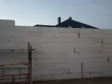 Budowa domu w toku. Ściany z bloczków betonowych czekają na tynkowanie. W tle widać dach i kominy.