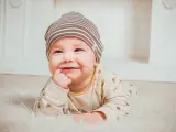 Uśmiechnięte dziecko w czapce leży na białym futerku, wkładając palec do buzi.