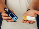 Man holds a vape in one hand and cigarettes in the other, posing the question: que es mejor el vaper o el tabaco.