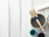 Herramientas para pintar mesitas de noche: lata de pintura gris, pincel, rodillo y botes de pintura azul y negra sobre fondo de madera blanca.