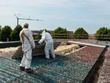 Dwóch pracowników w kombinezonach ochronnych usuwa stare płyty eternitowe z dachu.