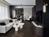 Nowoczesny salon z wygodną sofą, stolikami i regałem pełnym książek. Dominują ciemne, grafitowe akcenty, tworząc elegancką atmosferę.