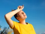 Une jeune femme en t-shirt jaune, le visage marqué par la chaleur, essuie son front sous un ciel bleu intense. La durée d'une insolation est palpable.