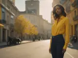 Une femme porte une chemise jaune oversize rentrée dans un jean. Elle se tient dans une rue bordée de bâtiments jaunes et d'arbres aux feuilles d'automne.