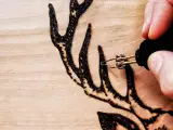 Dettaglio di un disegno a pirografia su legno: una mano guida le punte pirografo per creare le corna di un cervo.