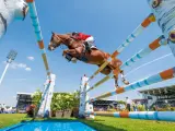 Un magnifique cheval de course brun saute par-dessus un obstacle coloré, sous un ciel bleu clair, lors d'une compétition équestre devant une foule attentive.