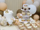 Biały tort z złotym misiem, babeczki i makaroniki na paterze, otoczone balonami w pastelowych kolorach.