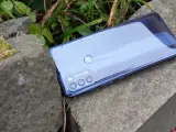 Błyszczący tył telefonu **Moto G 50** z potrójnym aparatem, leżący na kamieniach wśród zieleni.