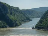 Szeroka rzeka przepływająca przez skaliste, zalesione góry. Widoczne łodzie i budynek.