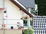 Dwóch mężczyzn na rusztowaniu maluje białą elewację domu. Sprawdź, jaka jest malowanie elewacji cena.