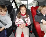 Tres niños sonríen en sus asientos de coche. El niño de la izquierda, con sudadera gris, parece preguntarse cuándo puede ir un niño delante.