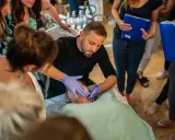 Mężczyzna w rękawiczkach demonstruje masaż kobido na twarzy leżącej osoby. Wokół zgromadzone kobiety obserwują.