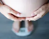 Dłonie obejmujące brzuch ciężarnej, stojącej na wadze. Schudłam w ciąży 10 kg, co widać na wadze.