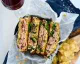 En saftig reuben sandwich, skuren i fyra bitar och toppad med färsk persilja. Serveras med krispiga potatischips och en glas rödvin.