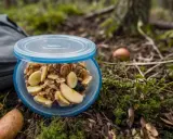 Idealne co do jedzenia w góry: musli z bananem i jabłkiem w pojemniku, obok plecak i jabłka na leśnym poszyciu.